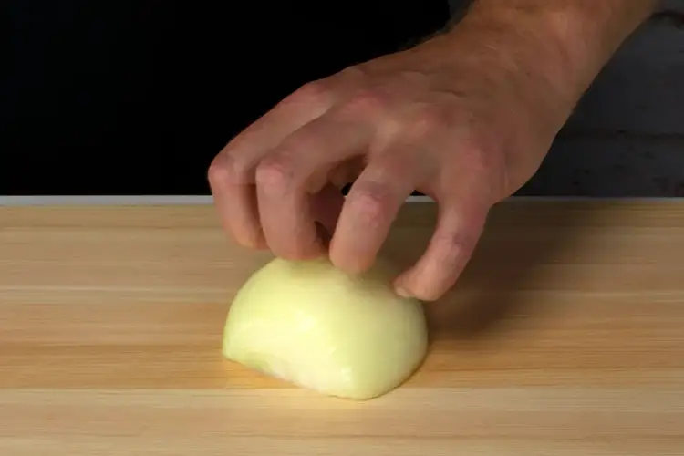 Hand demonstrating the claw grip technique while holding half an onion on a hinoki cutting board.